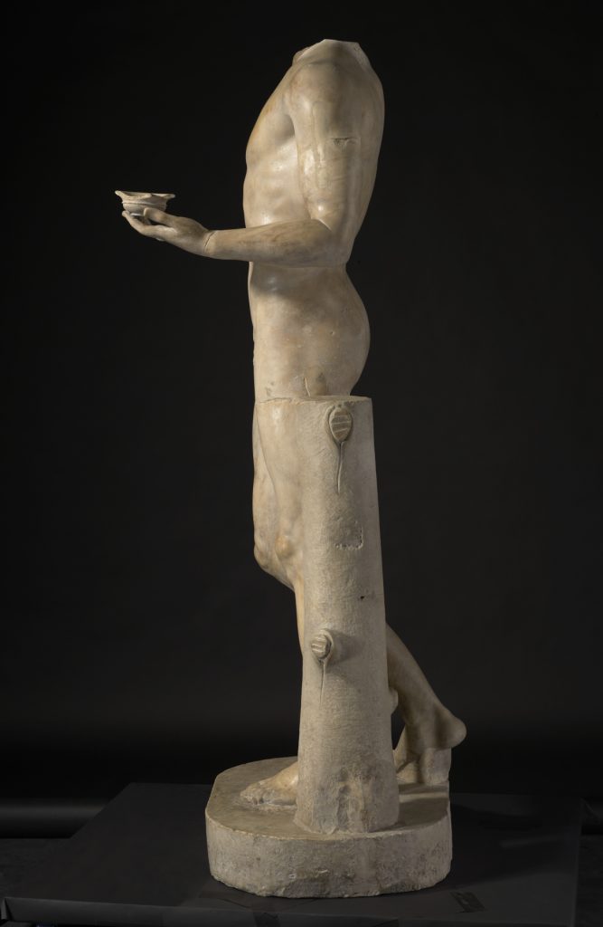 A white marble statue of a nude male figure wearing a crown and holding a bunch of grapes above his head with one hand and a cup in the other.