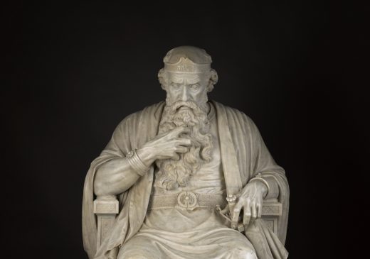 A life-sized white marble sculpture of King Saul seated on his throne against a black background. He wears robes, sandals, a belt, and a crown. His right hand clutches his long beard, and his left hand touches the handle of his dagger. His brow is furrowed.