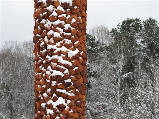 A tall, textured, red-orange sculpture on a grassy hill, with tall trees and a blue sky in the background. The sculpture has a cylinder shape that tapers to a point at the top and the bottom.