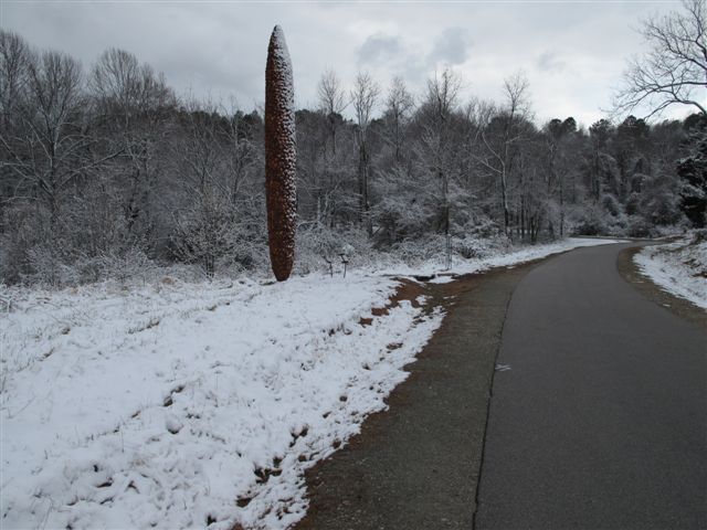 A tall, textured, red-orange sculpture on a grassy hill, with tall trees and a blue sky in the background. The sculpture has a cylinder shape that tapers to a point at the top and the bottom.