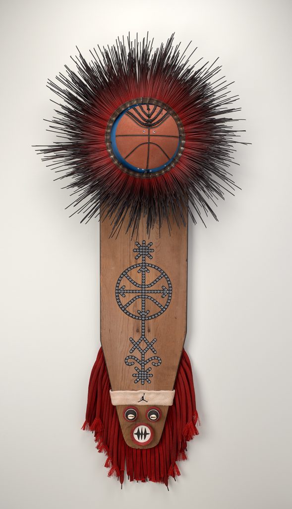 A wooden ironing board decorated with cowrie shells, braided synthetic hair, shoe laces, miniature clay pots, and a basketball.
