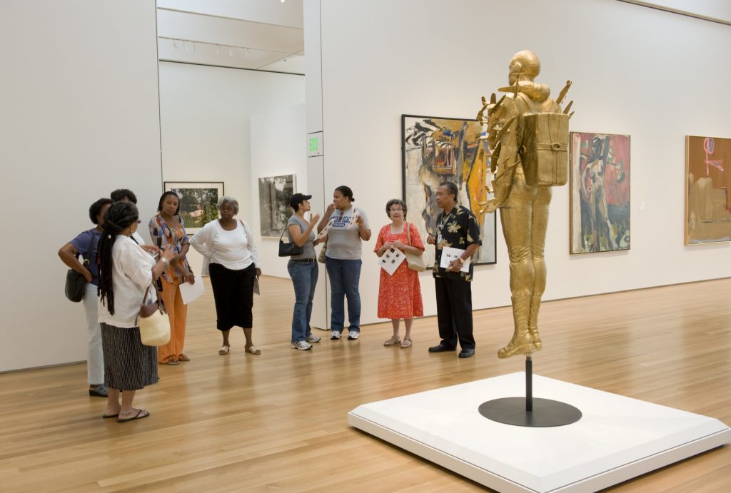 A gold-painted, life-size resin cast of Michael Richard’s full body, dressed in a Tuskegee Airman uniform, with dozens of small airplanes crashing into his arms and torso. The sculpture is mounted on a black metal stand that makes it appear to be suspended in the air.