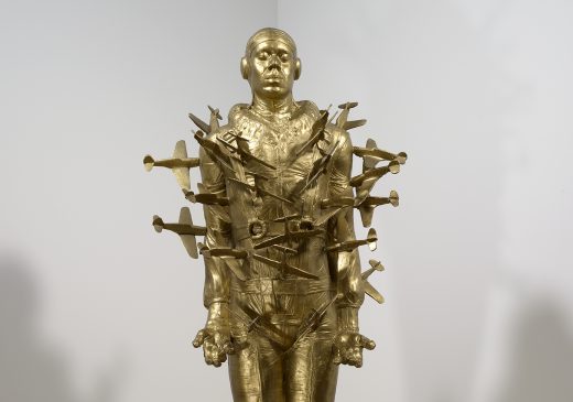 A gold-painted, life-size resin cast of Michael Richard’s full body, dressed in a Tuskegee Airman uniform, with dozens of small airplanes crashing into his arms and torso. The sculpture is mounted on a black metal stand that makes it appear to be suspended in the air.