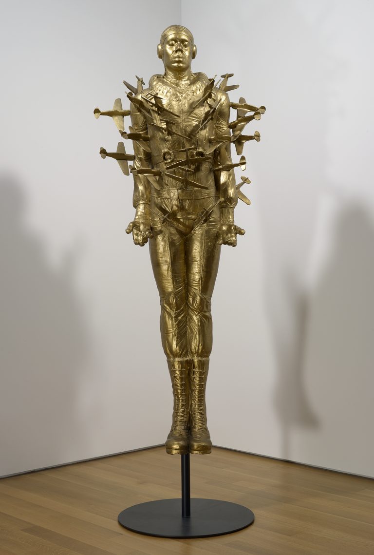 Un molde de resina de tamaño natural y pintado en oro del cuerpo completo de Michael Richard, vestido con el uniforme de aviador de Tuskegee, con docenas de pequeños aviones que se estrellan contra sus brazos y su torso. La escultura está montada en un soporte de metal negro que hace que parezca estar suspendida en el aire.