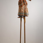 A sculpture of a headless female figure wearing a Victorian-era dress with a Dutch wax print design. Her arms are outstretched on each side of her body, palms facing up, and the figure is balanced on top of a pair of six-foot-tall wooden stilts.