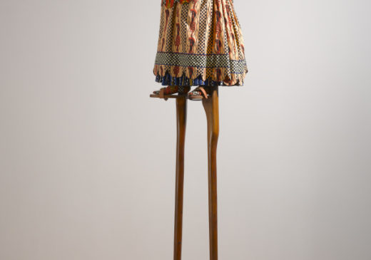 A sculpture of a headless female figure wearing a Victorian-era dress with a Dutch wax print design. Her arms are outstretched on each side of her body, palms facing up, and the figure is balanced on top of a pair of six-foot-tall wooden stilts.