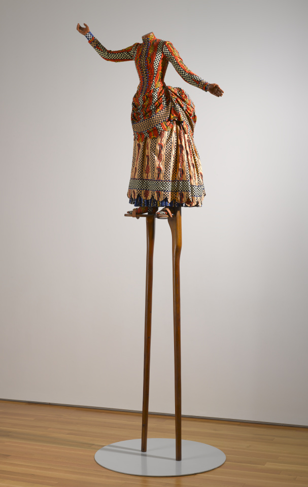 A sculpture of a headless female figure wearing a Victorian-era dress with a Dutch wax print design. Her arms are outstretched on each side of her body, palms facing up, and the figure is balanced on top of a pair of six-foot-tall wooden stilts.