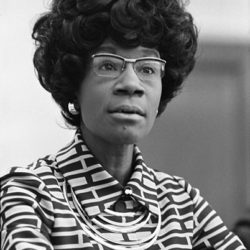 black and white photo of politician Shirley Chisholm wearing glasses and a high contrast collared shirt with a rectangle pattern
