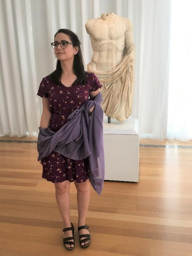 A woman in a purple dress holds a draped purple cloth in front of a marble sculpture.