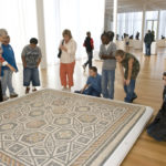 Group of people look at the mosaic floor displayed on the floor inside the galleries.