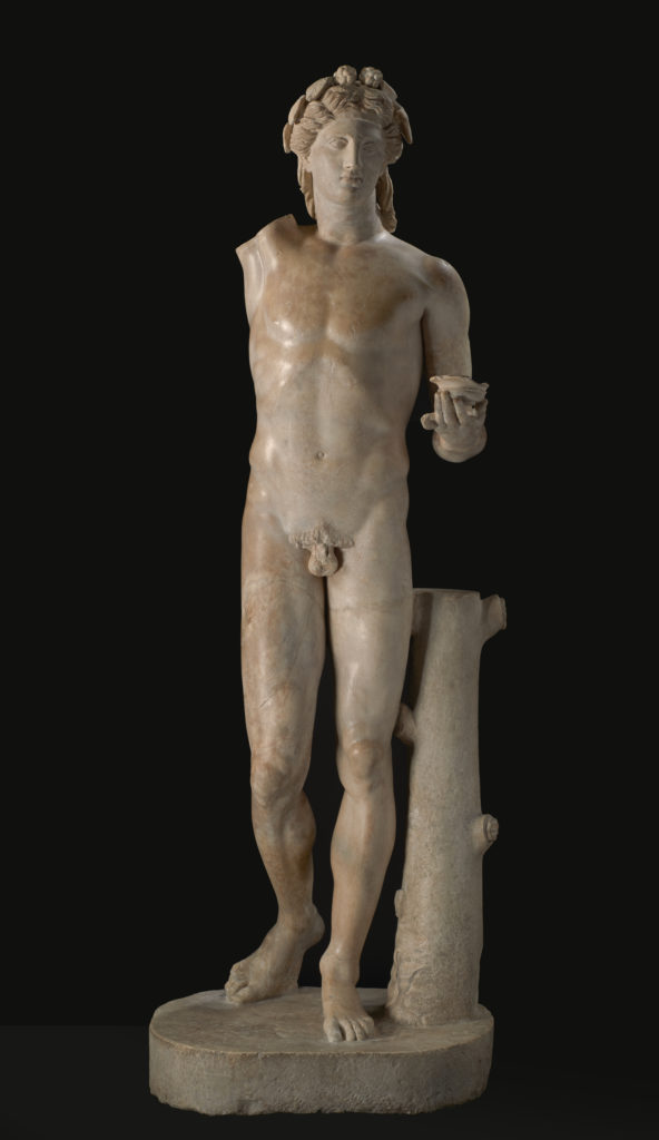 Estatua de mármol blanco de una figura masculina desnuda que lleva una corona y sostiene un racimo de uvas sobre su cabeza con una mano y una copa en la otra.