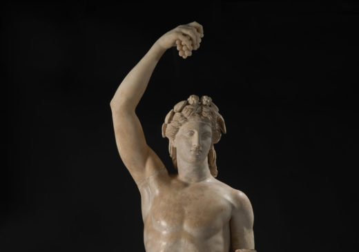 A white marble statue of a nude male figure wearing a crown and holding a bunch of grapes above his head with one hand and a cup in the other.