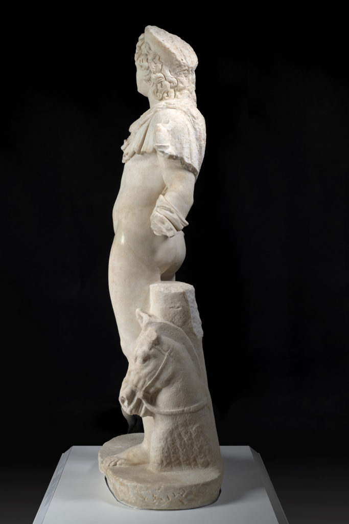 A marble statue of the Greek sun god Helios, without arms, depicted wearing only a short cloak over one shoulder and a partial crown, with the head of a horse at his feet.