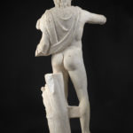 A marble statue of the Greek sun god Helios, without arms, depicted wearing only a short cloak over one shoulder and a partial crown, with the head of a horse at his feet. View of reverse.