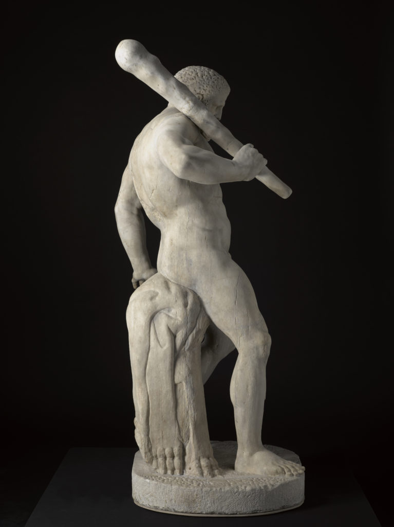 A white marble sculpture of a nude, bearded male figure holding a club over his shoulder. His weight is supported by a tree trunk behind him.