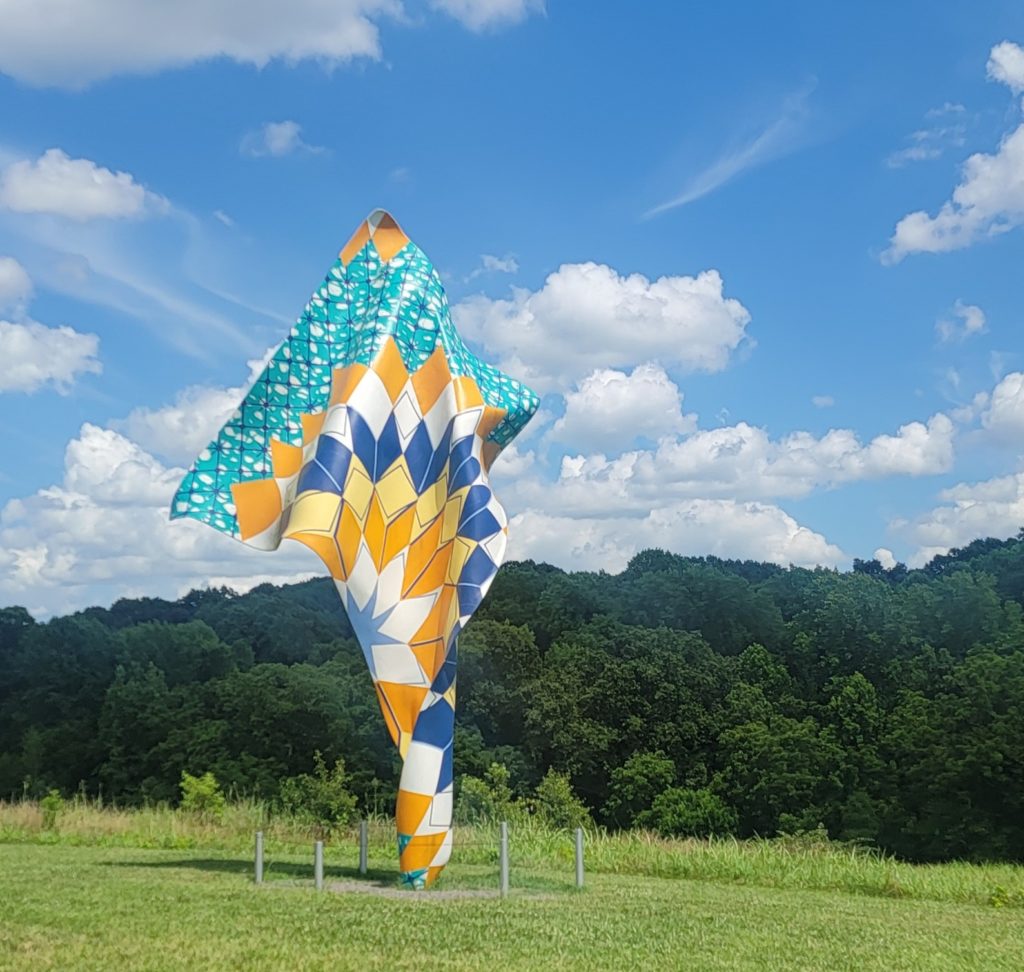 Una gran escultura de exterior que se asemeja a una tela fluida con diseños geométricos en tonos azules, amarillos, naranjas y blancos.