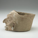 Side view of a light tan clay jar with a humanlike wrinkled face.