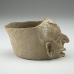Side view of a light tan clay jar with a humanlike wrinkled face.