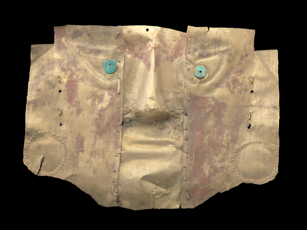 Una máscara de metal dorado con un rostro de aspecto humano, con ojos azules y verdes y vetas de pintura roja en las mejillas y la frente.