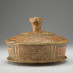 Front view of a terracotta cylindrical dish with a lid that has a jaguar-head handle.