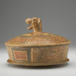 Side view of a terracotta cylindrical dish with a lid that has a jaguar-head handle.