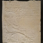Carved limestone relief sculpture with jagged top edge, showing a seated man facing right, surrounded by hieroglyphs and small offering figures. 