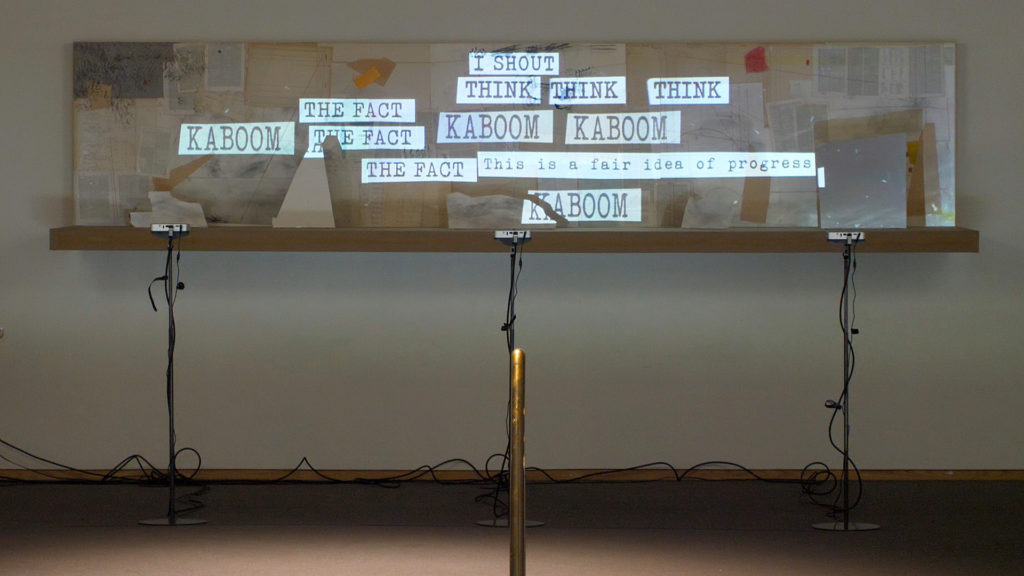 A photograph of an indoor film installation that includes three mini-projectors with stands. The following words and phrases are projected on a narrow, horizontal screen: I SHOUT, THINK, THE FACT, and KABOOM, and This is a fair idea of progress.
