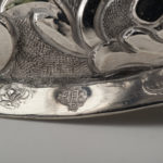 Detail view of the Kiddush Goblet base, showing a hand pouring a pitcher, with another hand supporting a basin below the pitcher.