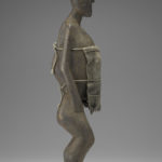 A small wooden sculpture of a woman with most of her torso covered by a burlap cloth. Side view.