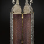 A hinged wooden case with domed finials on top and metallic floral patterns on the outside surface. View of opened case.