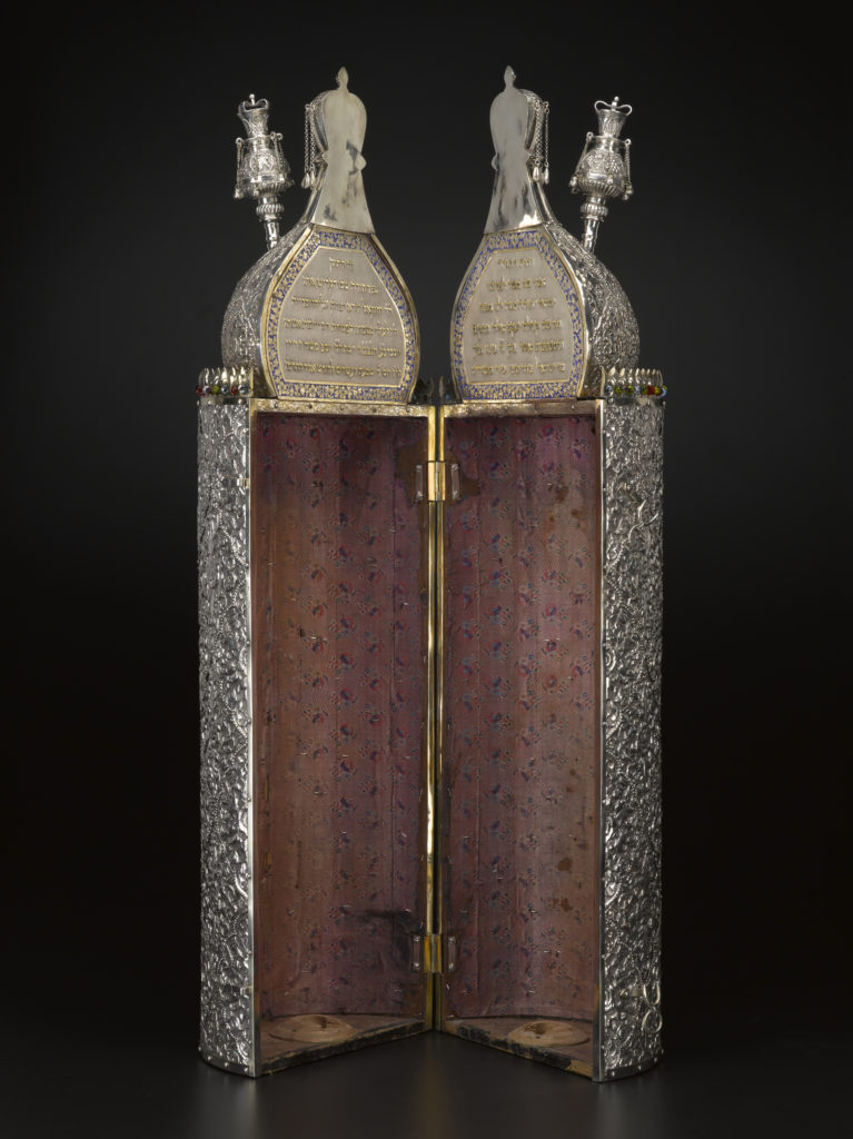 A hinged wooden case with domed finials on top and metallic floral patterns on the outside surface.