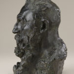 A bronze sculpture of the head, neck, and upper shoulders of a bearded man. Side view.