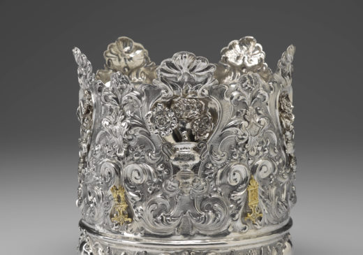 A silver crown covered with intricate details, including floral designs and Hebrew text.