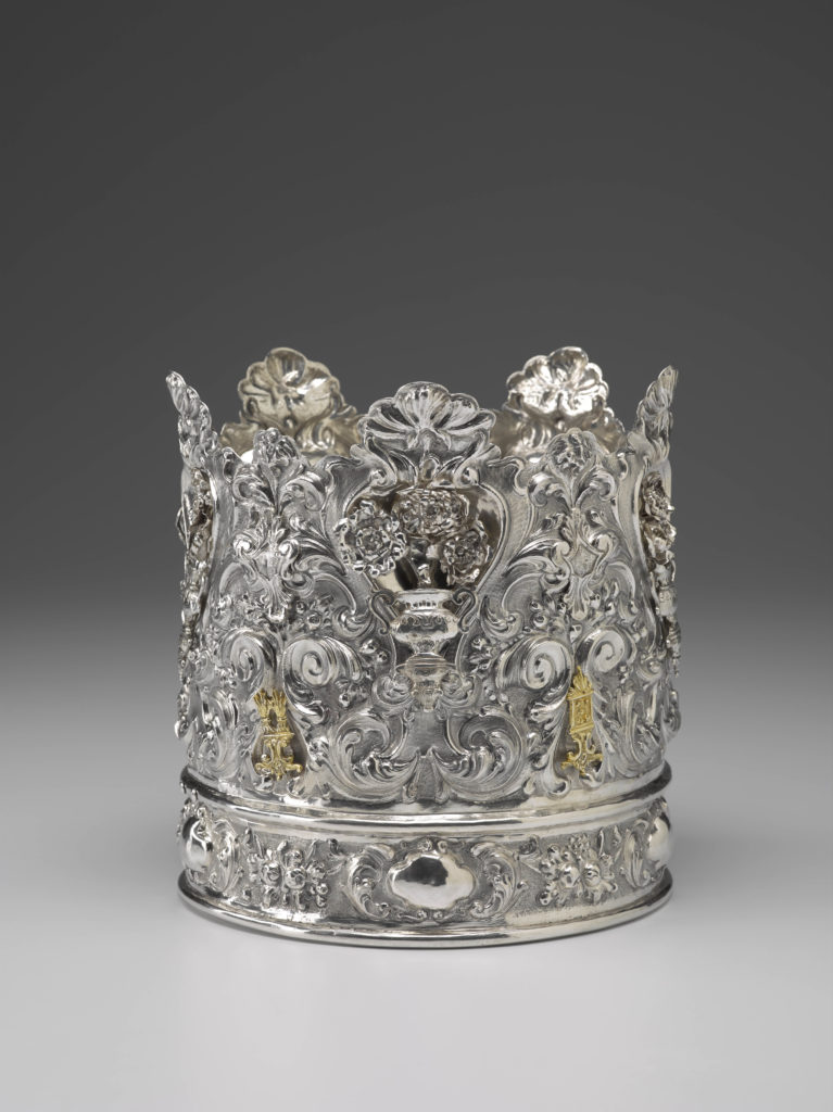 A silver crown covered with intricate details, including floral designs and Hebrew text.