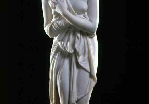 A white marble sculpture of a female figure. She is holding draped fabric that partially covers her nude body, and she is looking over her shoulder.