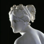 A white marble sculpture of a female figure. She is holding draped fabric that partially covers her nude body, and she is looking over her shoulder. Detailed view of face.