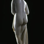 A white marble sculpture of a female figure. She is holding draped fabric that partially covers her nude body, and she is looking over her shoulder. View of the backside of the statue.