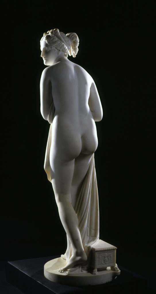 A white marble sculpture of a female figure. She is holding draped fabric that partially covers her nude body, and she is looking over her shoulder.