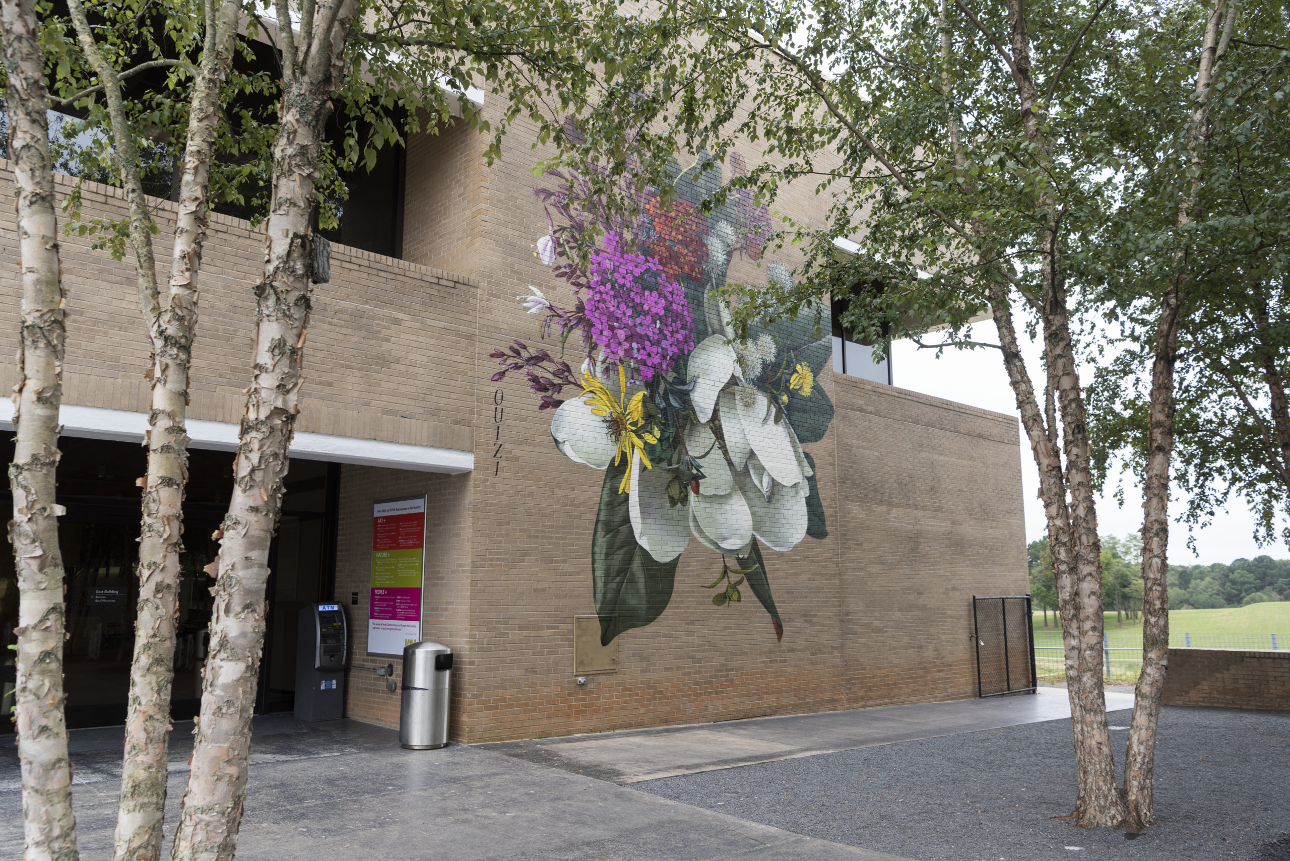 Summer’s Where You’ll Find Me A large-scale mural of white, yellow, and pink flowers painted on a brick building.