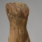 A wooden statue of a standing nude man with no arms, head, or neck. There are patches of brownish-red paint on the figure’s chest and legs.
