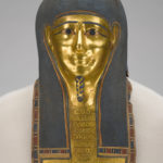 A white, linen-wrapped mummy covering with a gold Egyptian mask and gold accents on the chest, legs, and feet. Close up of funerary mask.