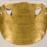 A white, linen-wrapped mummy covering with a gold Egyptian mask and gold accents on the chest, legs, and feet. Close up of breastplate.