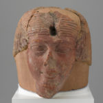 A brownish-red sculpture of a man’s head with a broken nose, a hole in the forehead, a partial beard, and a wig.
