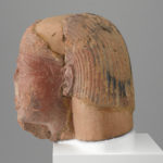 A brownish-red sculpture of a man’s head with a broken nose, a hole in the forehead, a partial beard, and a wig. Side view.