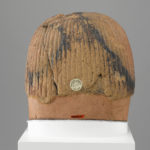A brownish-red sculpture of a man’s head with a broken nose, a hole in the forehead, a partial beard, and a wig. View of back.