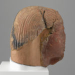 A brownish-red sculpture of a man’s head with a broken nose, a hole in the forehead, a partial beard, and a wig. Side view.