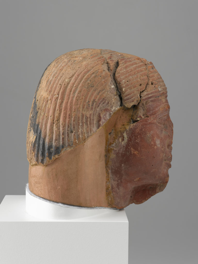 A brownish-red sculpture of a man’s head with a broken nose, a hole in the forehead, a partial beard, and a wig.
