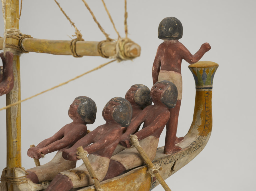 A wooden model of a boat with a crew of human figures and a passenger dressed in white clothing.