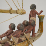 A wooden model of a boat with a crew of human figures and a passenger dressed in white clothing. Alternate view.