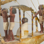 A wooden model of a boat with a crew of human figures and a passenger dressed in white clothing. Close up of figure in white clothing.